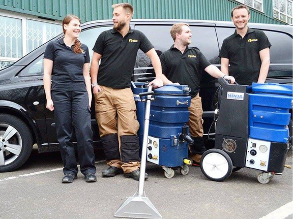 This is a photo of Belsize Park Carpet Cleaning Solutions carpet cleaners (three men and one woman) standing in fromt of their black van, with two steam cleaning carpet machines next to them